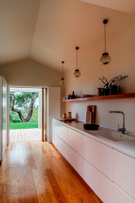 Small Iberian kitchen with white matte handleless cabinets running along one wall, white matte stone counter with single integrated sink and slim tap, a single floating warm-wood open shelf above the counter, three slim dark pendants hanging from a sloped plastered ceiling, wide-plank original pine floor, sliding door open to a sunlit deck with mature trees outside