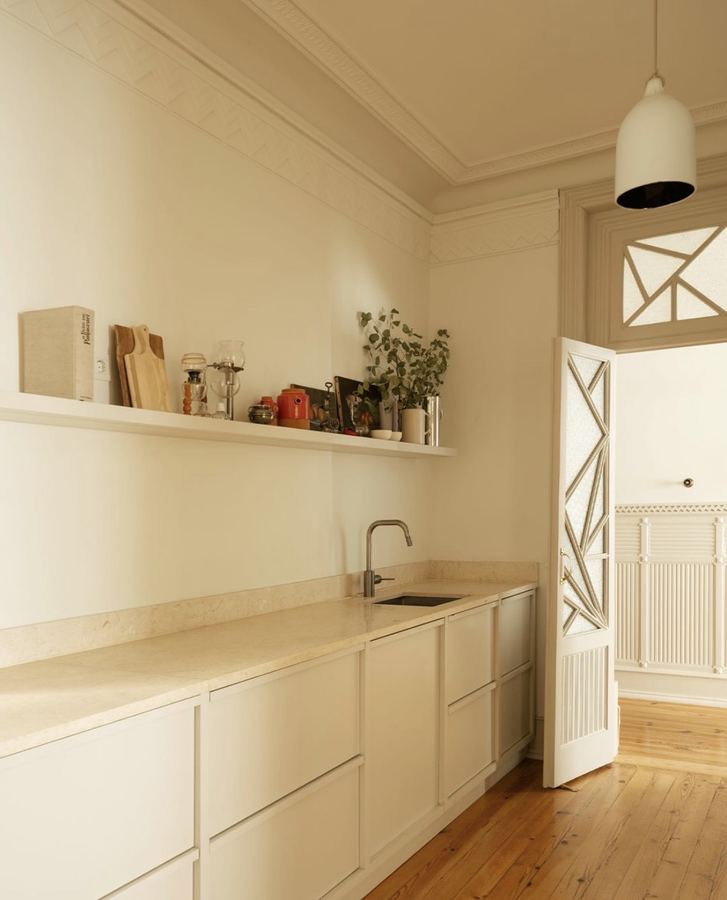 Cream-monochromatic kitchen with continuous cream stone counter, cream cabinets, single floating open shelf, traditional Portuguese door, ornate ceiling cornice, reclaimed pine floor