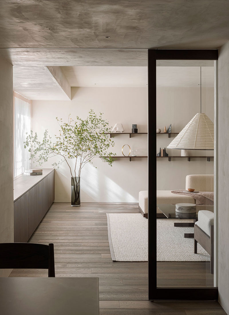 Norm Architects Karimoku Case Study Azabu Residence Tokyo interior: foreground room with raw plaster ceiling and warm off-white plaster walls, long pale light-oak low credenza along the left wall, tall slender glass vase with a delicate flowering branch on top, slim wall-mounted shelf above with small ceramic objects and a paper lampshade, sheer-curtained window on the left, a tall floor-to-ceiling slim black-framed sliding door at the right with translucent glass infill, beyond the door a living room with cream bouclé modular sofa, light-oak travertine-topped low coffee table on a pale wool rug, cream pleated origami fan pendant lamp, wall-mounted dark wood shelves with display objects, light-oak parquet floor