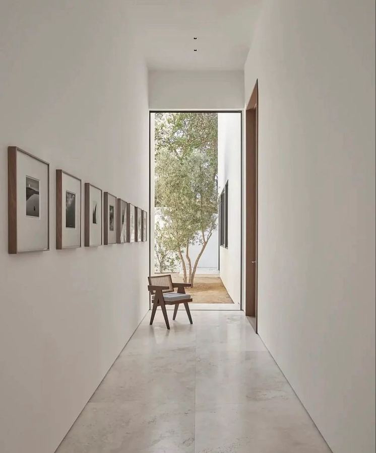 Long white-plaster corridor with a line of dark-framed photographs, a Pierre Jeanneret-style chair at the end as a sculptural focal point, large window onto an olive tree, dark wood door on the right