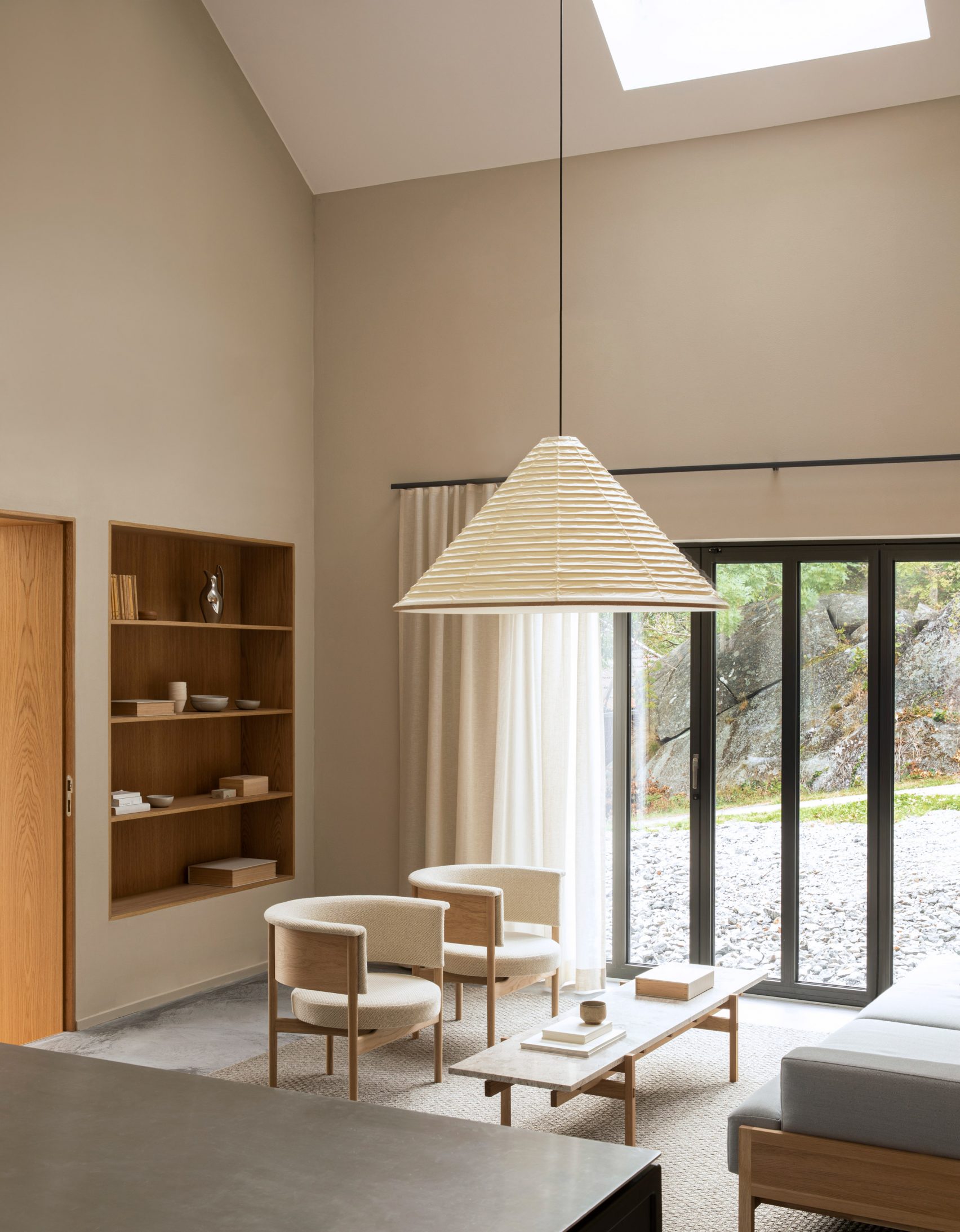 Norm Architects Archipelago House Sweden interior: high cathedral / sloped white ceiling with a skylight, warm ochre-beige plaster walls, recessed built-in light-oak open shelving with horizontal planks, light-oak flush door, two cream bouclé curved barrel armchairs, simple light-oak coffee table with travertine top, large cream pleated origami fan pendant lamp slung from the ceiling on a slim black cord, sisal seagrass area rug, polished concrete floor, slim black-framed sliding glass doors onto rocky archipelago landscape, cream linen floor-to-ceiling curtains