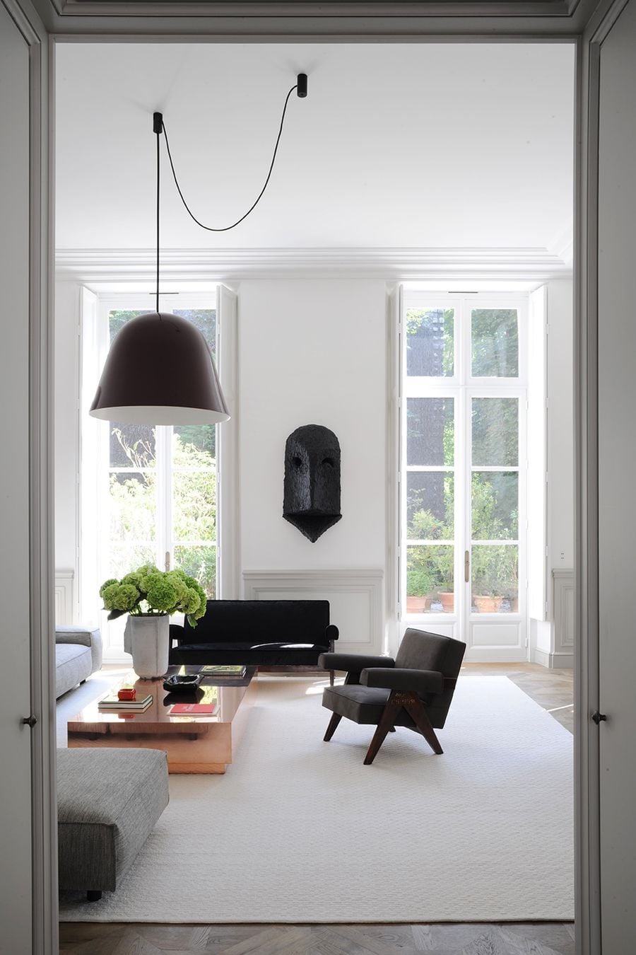 Joseph Dirand Saint-Germain Paris apartment: pure-white classical boiserie panelled walls with full-height divided-light French double doors onto a Parisian garden, ornate classical cornice and dado, light herringbone parquet floor, dark navy/midnight velvet sofa, Pierre Jeanneret-style upholstered grey lounge chair with dark wood frame, low rectangular coffee tables with white-marble top and bronze block base, oversized black Bouroullec Bells pendant lamp with exposed black cable swag from the ceiling, large dark sculptural African mask on the wall as the single artwork, tall white plaster vase with hydrangea, cream area rug, double-door portal frame in foreground