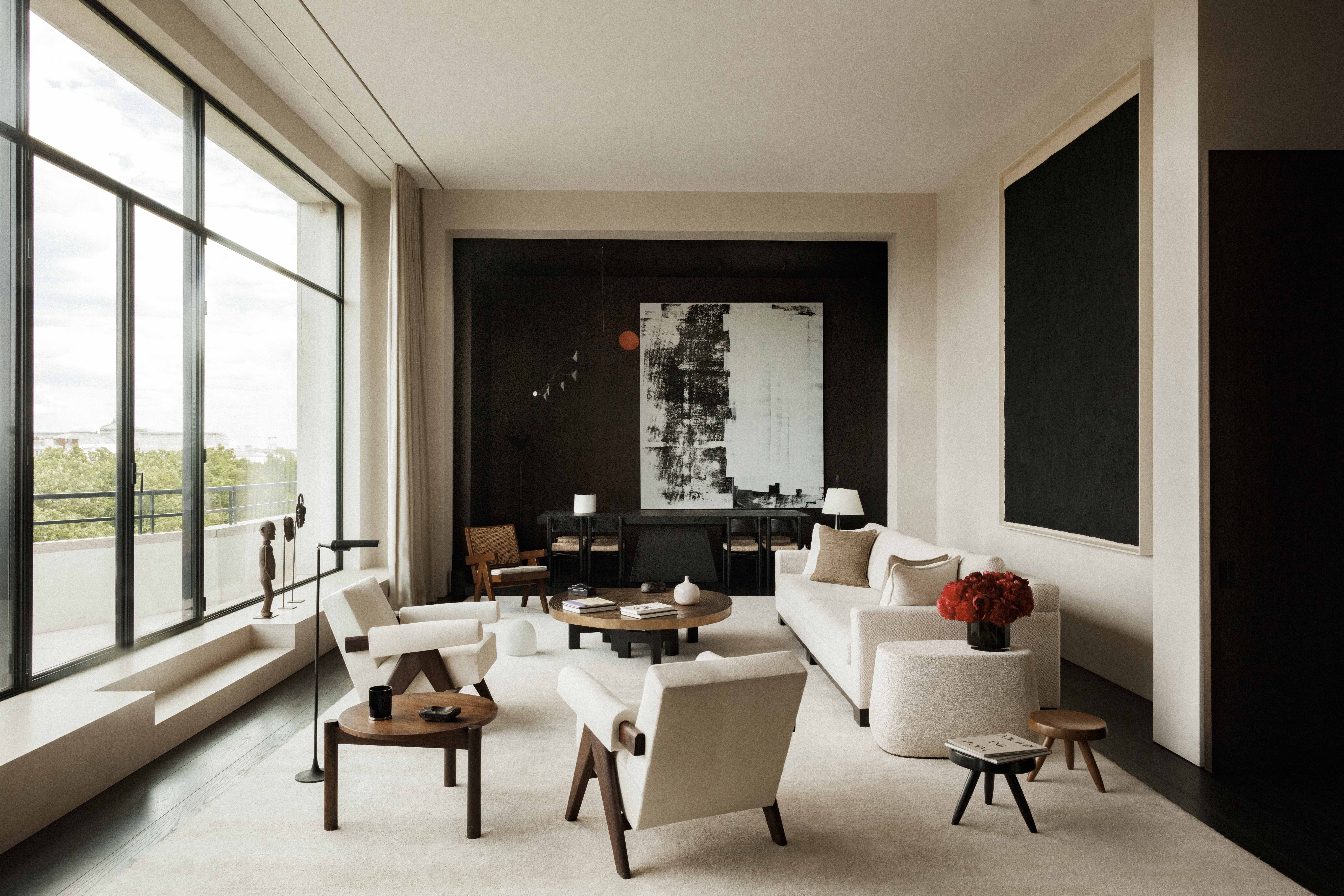 VVD RE Residence salon: tall Crittall-style slim black-steel floor-to-ceiling window onto Parisian rooftops, cream bouclé sofa and armchairs, dark walnut Pierre Jeanneret-style lounge chair, low round dark walnut coffee table, small black tripod stool, slim black floor lamp with cream shade, dark stained oak floor, pale sisal rug, dark accent wall in the back framing a large abstract black-and-white painting with a small orange dot, large dark monochromatic painting recessed in a cream-framed niche on the right, cream linen curtains, built-in plaster window bench