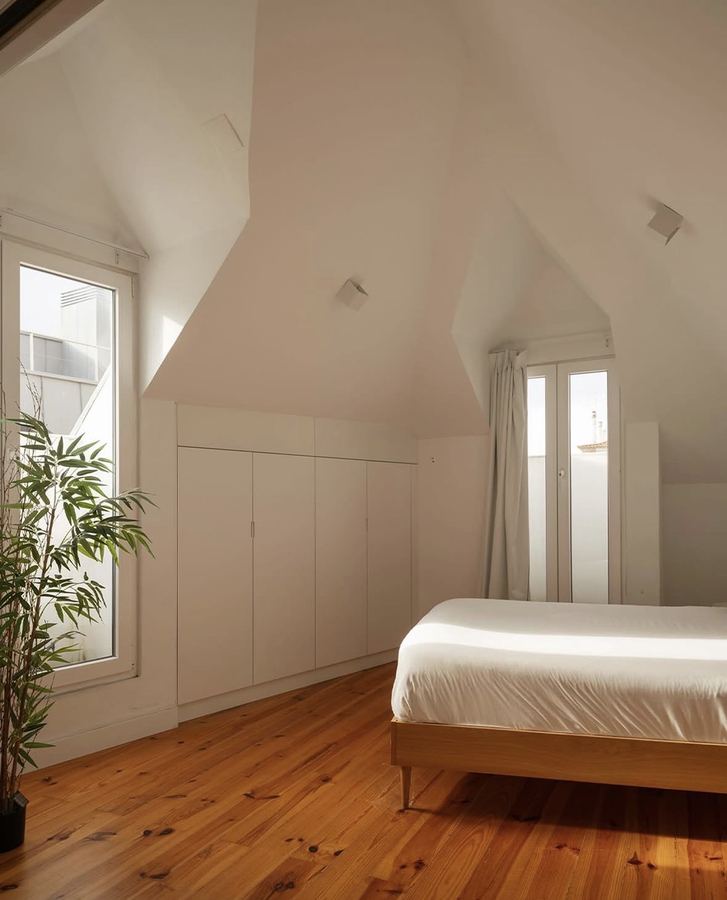 Minimal bedroom with white plaster, light wood plank floor preserved and lightened, simple low oak bed frame with white linens and no headboard, built-in white wardrobes with push-to-open fronts, narrow windows