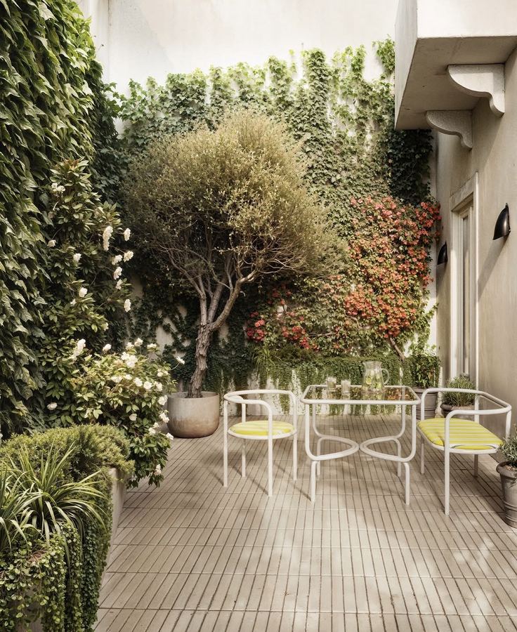 Lush courtyard with walls covered in dense ivy, flowering bushes (white roses, orange-pink azaleas), small olive-like tree in terracotta pot, vintage white-painted metal table and curved chairs with yellow striped seats, patterned cement-tile (azulejo hidráulico) floor, black wall sconce