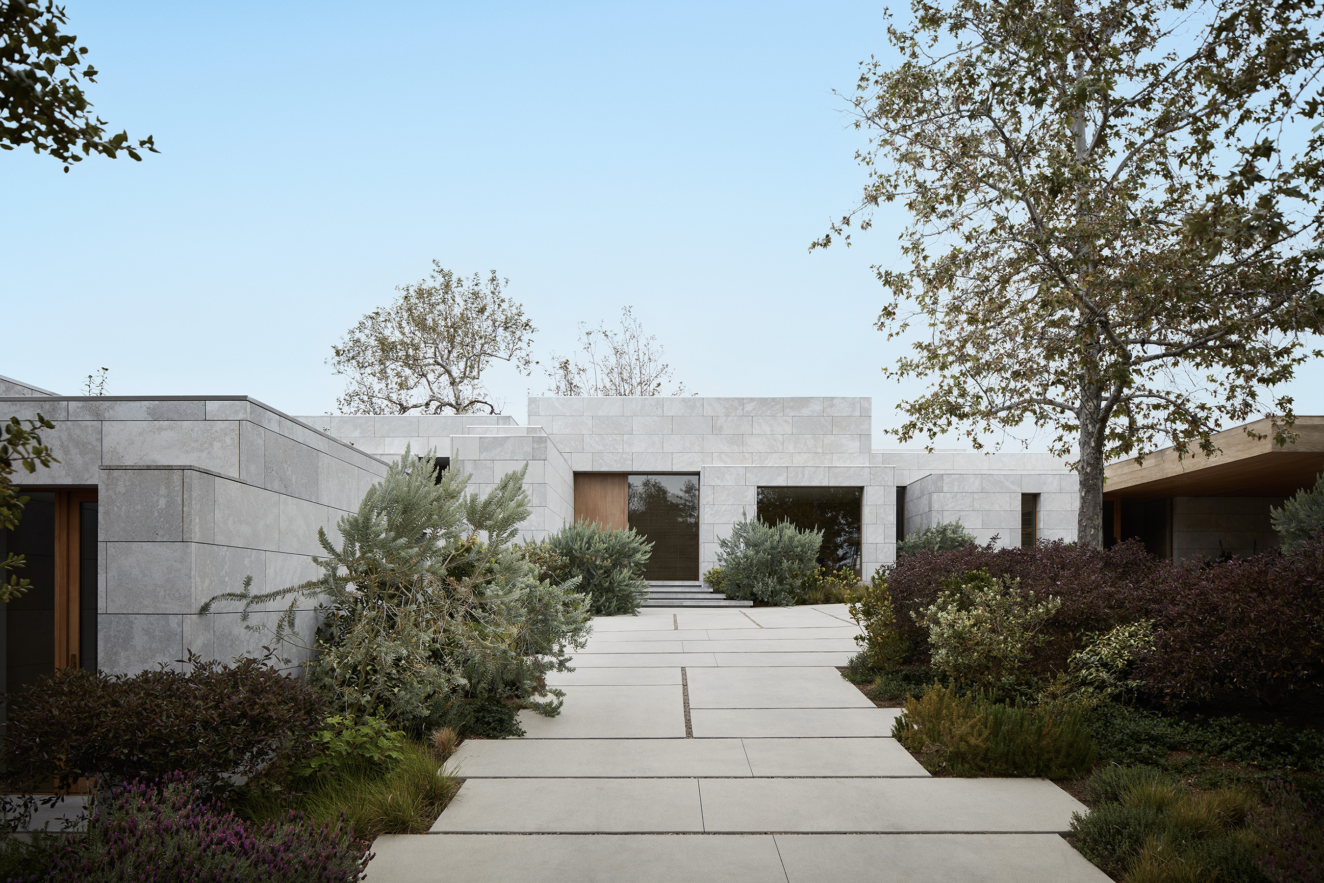 Vincent Van Duysen AN Residence exterior approach: low single-storey volume clad in pale grey limestone, walkway of large-format light grey concrete pavers with thin grout lines, Mediterranean planting (rosemary, lavender, ornamental grasses, succulents), mature trees, pale clear sky